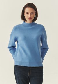 Pull bleu clair en maille avec un col montant, des poignets et un ourlet côtelés. Porté avec un jean foncé, avec une coupe décontractée et des manches longues.