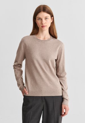 Femme aux longs cheveux châtain clair portant un pull beige à col rond et un pantalon gris foncé, debout avec une main dans la poche devant un fond gris.