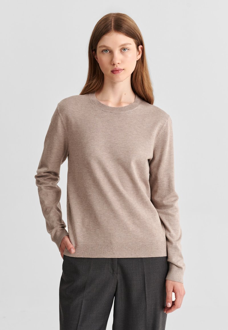 Femme aux longs cheveux châtain clair portant un pull beige à col rond et un pantalon gris foncé, debout avec une main dans la poche devant un fond gris.