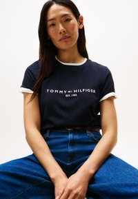 Młoda kobieta o długich ciemnych włosach w granatowej koszulce Tommy Hilfiger i niebieskich dżinsach, siedząca z rękami spoczywającymi na kolanach na jednolitym tle.