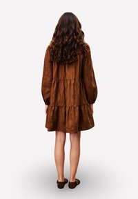 Frau mit langen, lockigen Haaren, die ein braunes, gestuftes Kleid und braune Ballerinas trägt, steht mit dem Rücken zu einer weißen Hintergrund.