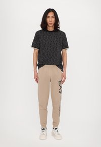 Svart mønstret t-skjorte med korte ermer, beige joggebukser med et grafisk design på siden, kombinert med hvite sneakers.