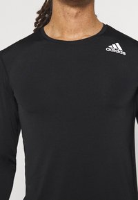 Person, der ein schwarzes Langarmshirt mit einem weißen Adidas-Logo auf der rechten Schulter trägt, zeigt Nacken und oberen Oberkörper.