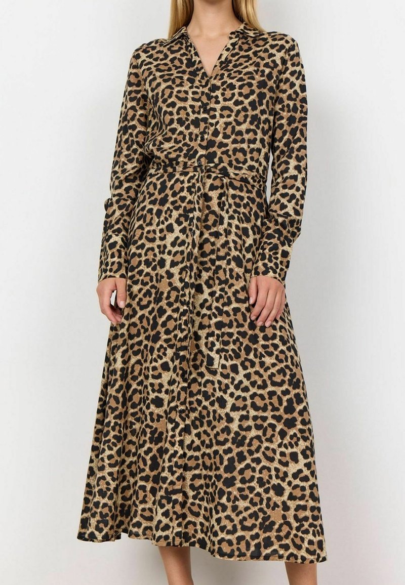 Femme portant une robe midi à manches longues boutonnée, imprimé léopard, avec ceinture nouée à la taille sur un fond uni.