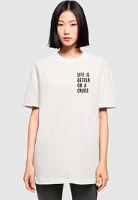 Weißes Baumwoll-T-Shirt mit kurzen Ärmeln, das auf der linken Seite schwarzen Text mit der Aufschrift "DAS LEBEN IST BESSER AUF EINEM KREUZFAHRT" zeigt.