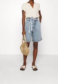 Světle béžová texturovaná blůza s krátkým rukávem, džíny s vysokým pasem a opaskem, tkaná slaměná taška a světlé sandály s detaily na přezce.