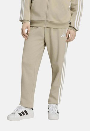Pantaloni da ginnastica beige Adidas con strisce bianche e logo, indossati con sneakers bianche con strisce nere.