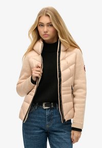 Padded jacket z miękkiej beżowej tkaniny z czarnymi akcentami. Wysoki kołnierz, zamek błyskawiczny i dwie boczne kieszenie. Nosi się go na czarnym swetrze.