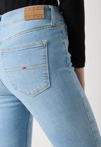Jean en denim bleu clair, de coupe ajustée. Présente une patch en cuir étiqueté "TOMMY JEANS" et un petit logo en rouge, blanc et bleu.