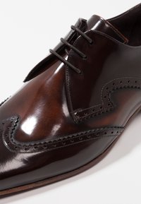 Chaussure habillée en cuir marron avec une finition polie, un design à bout golf, des perforations décoratives et une forme pointue à lacets.