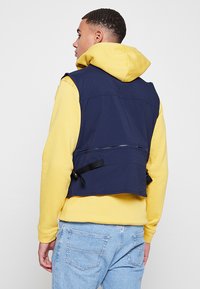 Gilet senza maniche blu navy con zip sul retro, cinghie laterali regolabili e tessuto liscio, indossato sopra una felpa gialla con cappuccio.