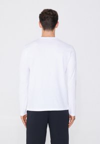 Langärmliges weißes Baumwoll-T-Shirt mit Rundhalsausschnitt, flachem Saum und minimalistischem Design, getragen mit navyblauen Hosen. Rückansicht dargestellt.