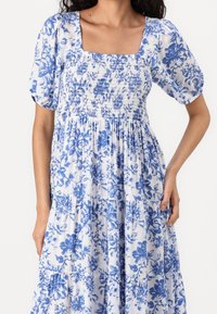 Robe longue florale bleue et blanche avec manches ballon, encolure carrée, corsage smocké et jupe à volants. Tissu léger et texturé.