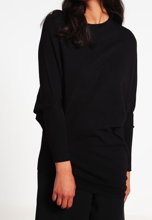 Femme portant un haut noir à manches longues, coupe décontractée, debout devant un fond blanc uni.