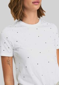 Camiseta blanca de manga corta con lentejuelas negras y plateadas dispuestas en un patrón disperso; textura suave y cuello redondo.