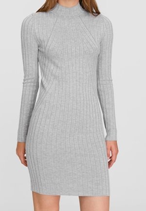 Femme portant une robe en maille gris clair, côtelée, à manches longues, longueur genou, avec un col montant.