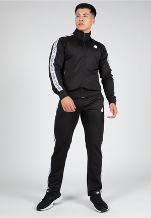 Zwart sportief trainingspak met ritsjack en bijpassende broek. Voorzien van contrasterende witte zijstrepen met tekst en een klein logo op het jack.