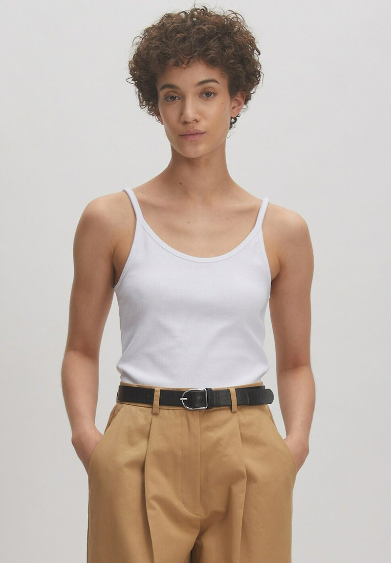 Witte katoenen tanktop met dunne schouderbanden, gecombineerd met hooghals beige broek en een zwarte riem met een zilveren gesp.