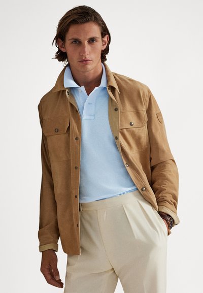 Béžový semišový kabát s dvoma náprsnými vreckami, nosený cez svetlomodrú polokošeľu. Kabát má zapínanie na patentky a štruktúrovaný povrch.