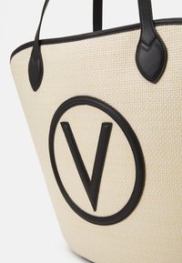 Tygväska med en krämfärgad vävd textur, som har ett stort svart lädercirkelsymbol med ett 'V' i mitten och svarta läderdetaljer.