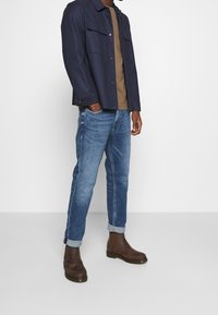 Chaqueta azul marino abotonada, camiseta marrón, vaqueros azules y botines marrones hasta el tobillo. La chaqueta tiene dos bolsillos en el pecho; los vaqueros son ligeramente ajustados con los puños doblados.