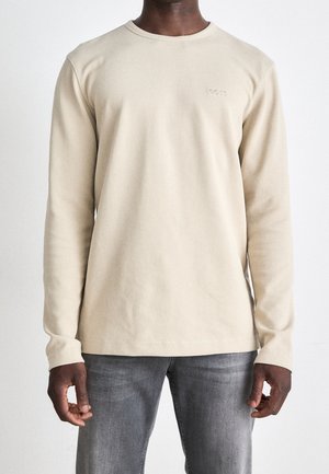 Mand iført beige langærmet ribstrikket skjorte med lille præget logo på brystet og grå denimjeans mod en ensfarvet baggrund.