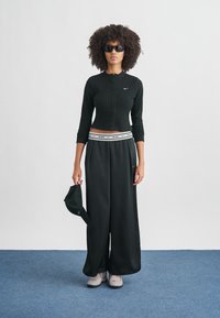 Μαύρο μακρυμάνικο τοπ με ρίγες και κουμπιά, άνετο μαύρο παντελόνι, γκρι παπούτσια και μαύρο καπέλο. Διαθέτει λογότυπο Nike στη μέση του παντελονιού και στο τοπ.