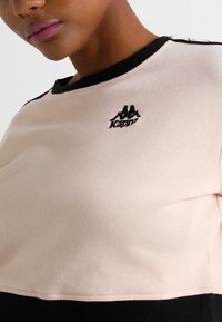 Primo piano di una persona che indossa una felpa Kappa beige e nera con logo sul petto e bordo nero sul collo e sulle spalle.