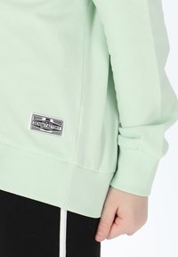 Felpa verde menta chiaro con orlo e polsini a costine. Presenta una toppa con logo in bianco e nero sul lato, con una mano visibile che la tiene parzialmente.