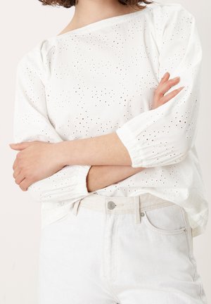 Frau trägt weiße Lochbluse mit Puffärmeln und helle Jeans mit hoher Taille, Arme vor der Brust verschränkt.