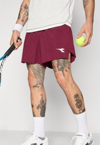 Pantalones cortos deportivos color marrón con un logo blanco, combinados con calcetines blancos y zapatillas. Sostiene una raqueta de tenis en una mano y una pelota de tenis verde en la otra.