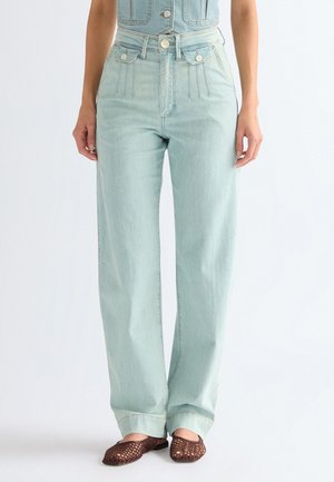 Lichtblauwe, recht gesneden jeans met hoge taille en gestikte voorzakken, gecombineerd met bruine geweven instapschoenen, gedragen door een staand persoon.