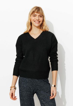 Pull noir à col en V côtelé avec manches longues, tissu texturé et taille ajustée. Porté avec un pantalon bleu marine à motifs et des accents de bijoux.