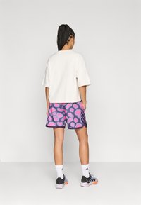 Κοντομάνικο κρεμ μπλουζάκι συνδυασμένο με μωβ σορτσάκια που έχουν ροζ αφηρημένα σχέδια. Το μοντέλο φοράει λευκές κάλτσες και μαύρα αθλητικά παπούτσια.