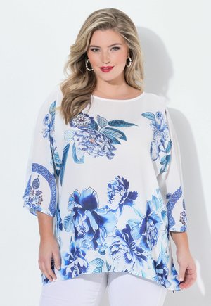 Frau mit welligem blondem Haar, die eine weiße Bluse mit blauen Blumenmustern und weiße Hose trägt, lächelnd vor einem einfarbigen Hintergrund.