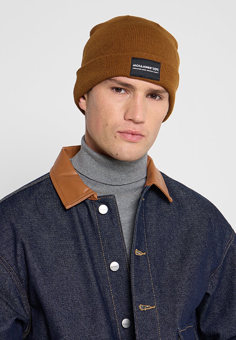 Bruine gebreide beanie met logolabel. Draagt een grijze coltrui en een blauwe spijkerjack met een beige leren kraag en metalen knopen.