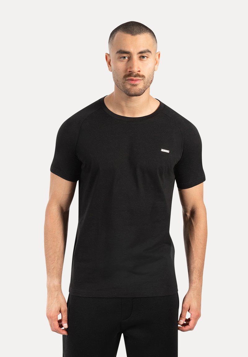 Man met kort haar en een getrimde baard, gekleed in een aansluitend zwart T-shirt, staand tegen een effen lichte achtergrond.