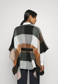 Poncho de cuadros en lana gris, negro, blanco y marrón. Presenta una cintura ajustada y una silueta fluida con una textura suave y mangas anchas.