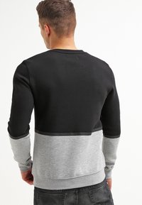 Hombre con una sudadera negra y gris de manga larga, visto desde atrás, con jeans oscuros sobre un fondo blanco sencillo.