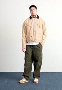 Chaqueta de corduroy beige con un cuello oscuro, camiseta blanca debajo, pantalones cargo oliva y zapatillas con acentos amarillos y un perfil bajo.