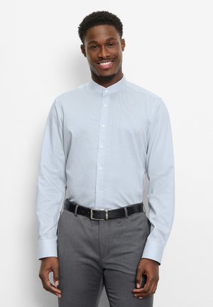 Homme souriant portant une chemise rayée bleu clair à col mandarin rentrée dans un pantalon gris avec une ceinture noire, debout devant un fond blanc.