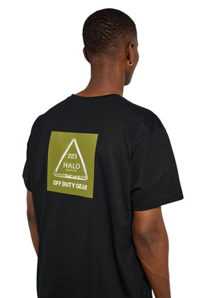 Persoon met kort haar die een zwart T-shirt draagt met een groen vierkant en witte tekst "223 HALO OFF DUTY GEAR" op de achterkant.