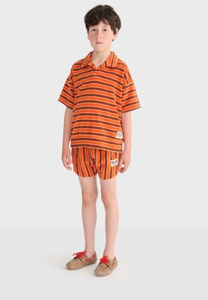 Dječak stoji, obučen u narančastu i smeđu prugastu majicu kratkih rukava i odgovarajuće kratke hlače, s bež cipelama i crvenim vezicama, na jednostavnoj svijetloj pozadini.