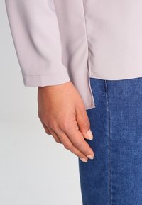 Blusa viola chiaro con maniche lunghe e una fessura laterale, abbinata a denim blu. Il tessuto appare liscio, con dettagli di cucitura visibili.
