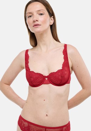 Femme portant un soutien-gorge en dentelle rouge et une culotte assortie, debout les mains derrière le dos sur un fond clair uni.