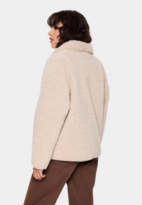 Beige fliist jaak, millel on tekstureeritud pind, kõrge krae ja relaxed fit. Kandmisel pruunide pükste kohal, näidates casual välimust.