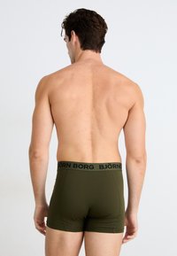Olivenfargede boxershorts med en kontrastfarget svart linning som har "BJÖRN BORG" i store bokstaver, laget av et glatt, stretchbart stoff.