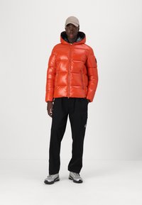 Bright orange puffer jacket with a zip front, elastic cuffs, and hood. Paired with black cargo pants and silver sneakers, set against a white background.