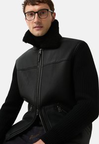 Zwarte leren vest met geribde gebreide mouwen en een hoge, gestructureerde kraag. Voorzien van een zilveren ritssluiting en twee zijzakken met ritssluiting.