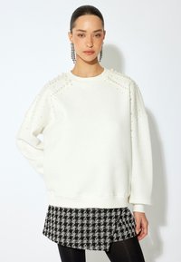 Sudadera blanca con adornos de perlas en los hombros y mangas largas, combinada con una falda de cuadros en blanco y negro, y leggings negros.